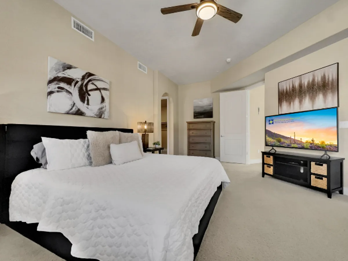 A tranquil bedroom with a neatly made bed, wall-mounted TV, and tasteful artwork above the headboard. The space features soft lighting and a calm, uncluttered atmosphere, inviting relaxation and privacy.
