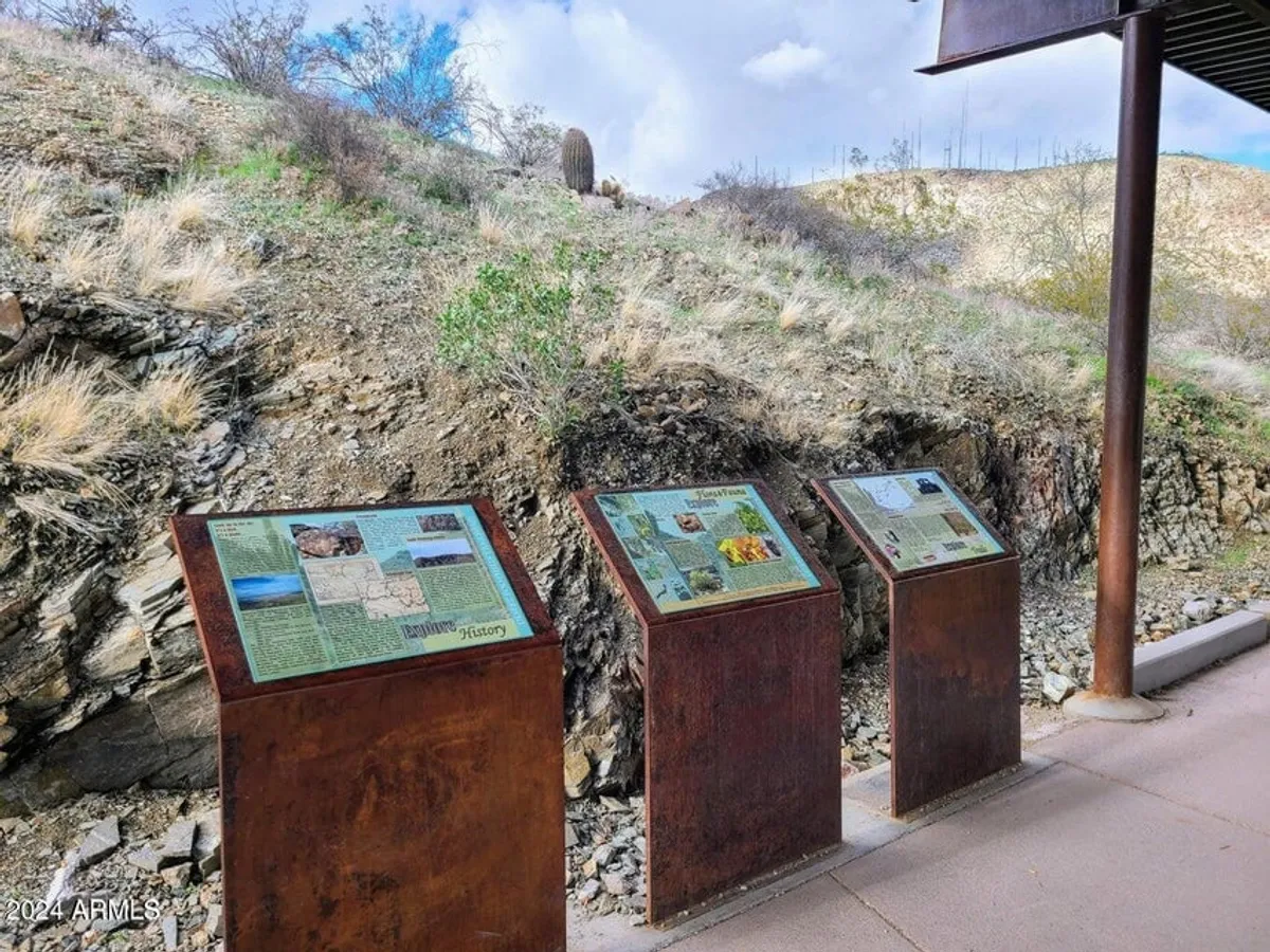 This image displays informational plaques set against a natural desert landscape with rocky terrain, encouraging exploration and engagement with the local environment. The scene is open and welcoming, inviting viewers to learn about the unique flora, fauna, and cultural history of the Phoenix area.