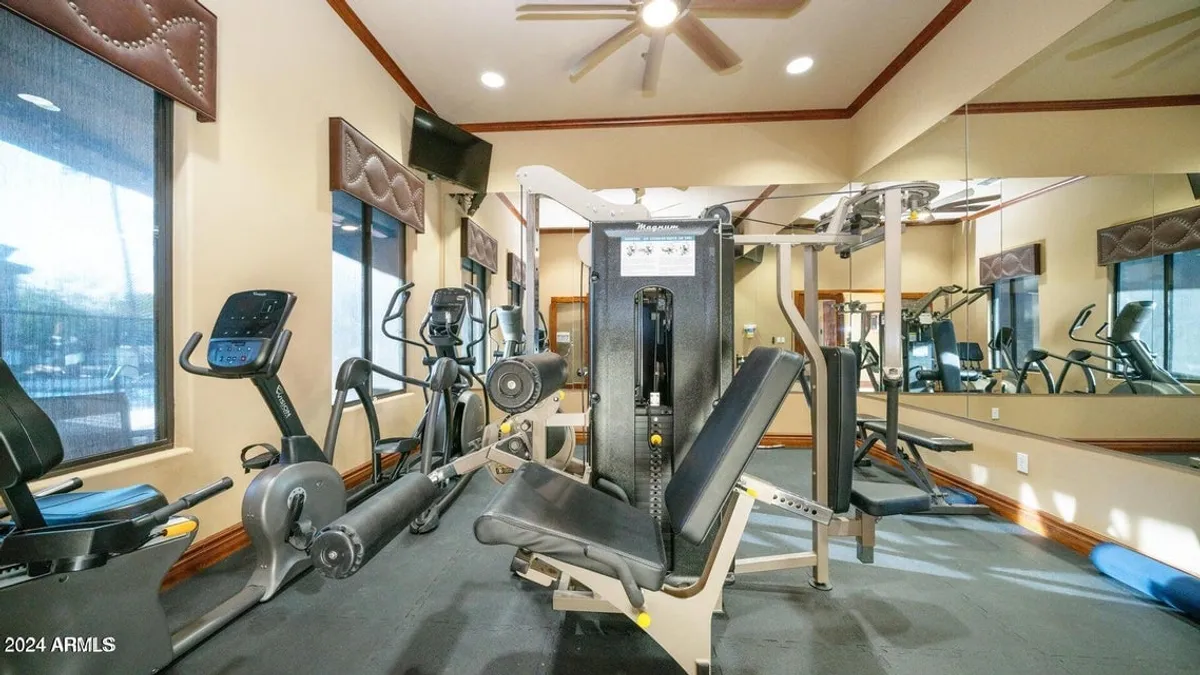 A bright, modern gym filled with exercise machines, mirrored walls, and natural light flooding in from the windows. The space is clean, well-equipped, and designed for both solo and community workouts.