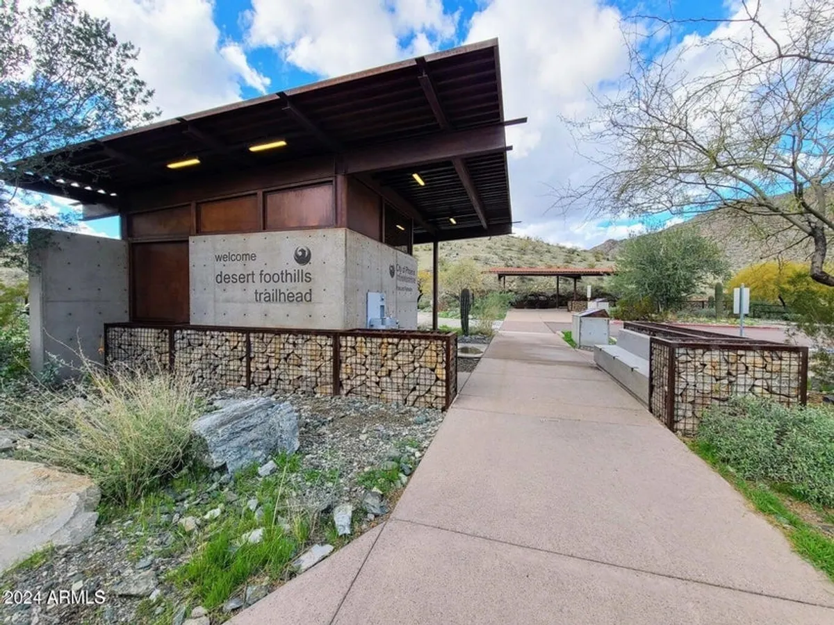 An inviting trailhead with a welcome sign and surrounding landscape, signaling the start of hiking or biking trails. The area feels accessible and ready for exploration, with hints of sunrise and wildflowers.