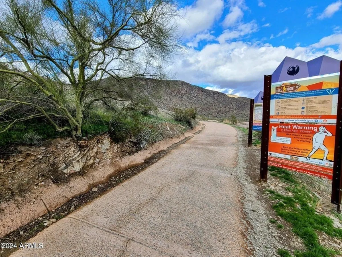 This exterior image features a walking path bordered by desert vegetation, an information sign, and a dramatic mountain backdrop, evoking the spirit of exploration and outdoor activity in the Ahwatukee area.