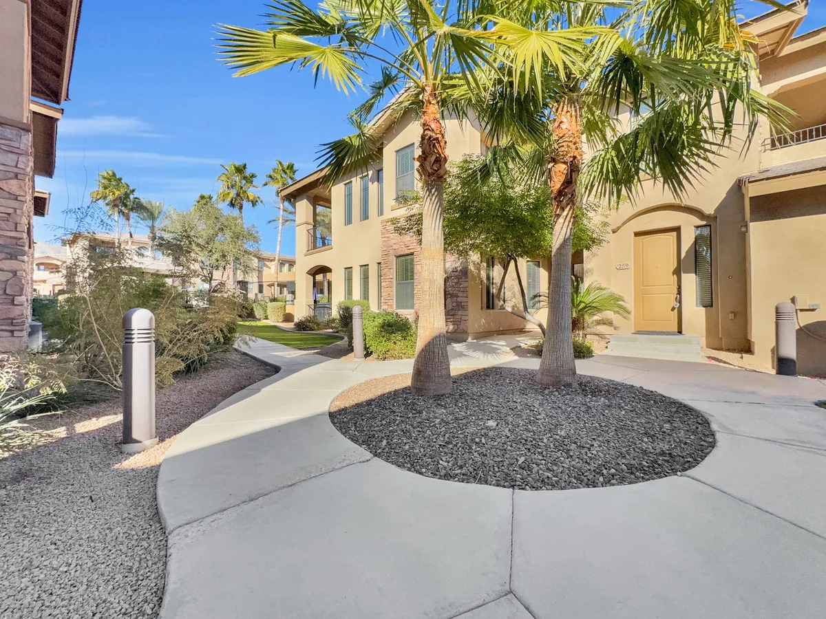 A welcoming exterior view featuring palm trees lining a curved pathway leading to the building's facade. The environment is lush and inviting, evoking a sense of daily life, relaxation, and connection with neighbors in a tranquil setting.