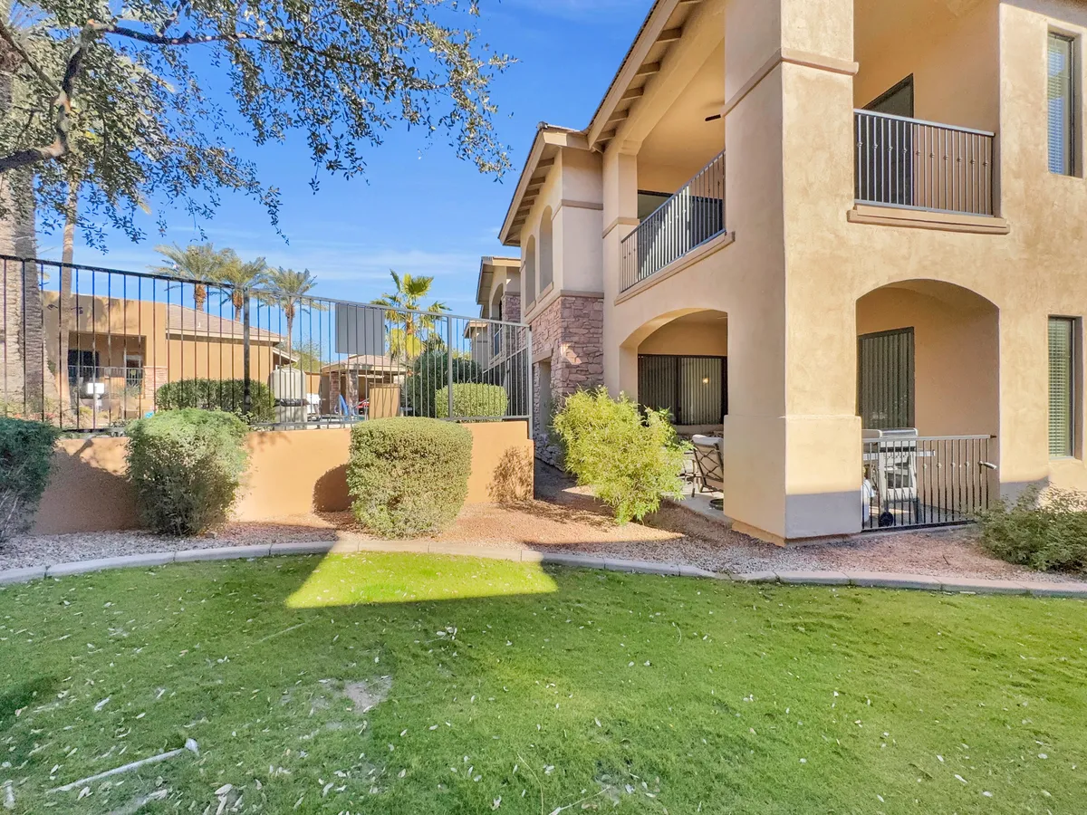 A tranquil exterior view of the condo complex, featuring a two-story building surrounded by a lush green lawn, manicured bushes, and balconies with railings. The setting is open, sunlit, and inviting, with ample outdoor space that could be enjoyed by both residents and their pets.