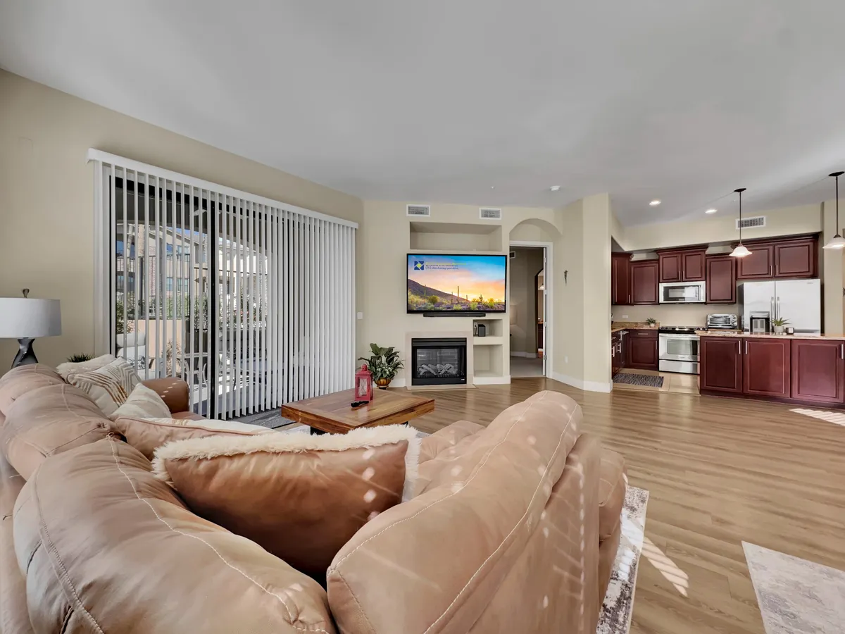 A spacious, open-plan interior featuring a large sectional sofa, a television, a fireplace, a visible kitchen area, and a dining table. The space feels inviting and functional, blending living, dining, and cooking zones seamlessly, and offers a view out towards a balcony for added openness.