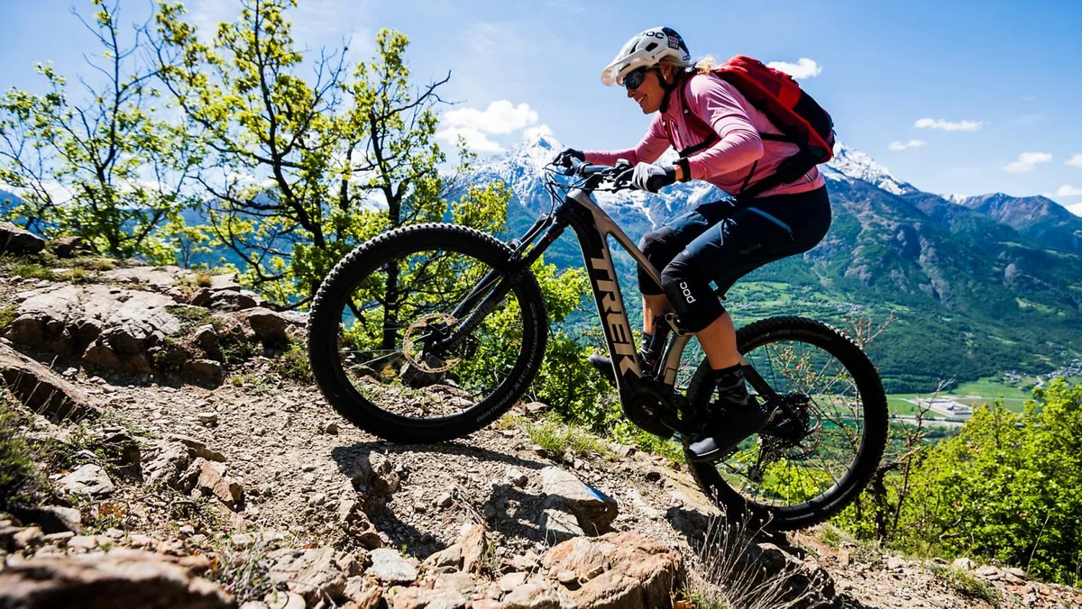 This exterior image captures a mountain biker navigating rocky terrain surrounded by distant mountains and lush greenery. The scene conveys excitement and adventure, with a focus on active outdoor pursuits in a natural setting.